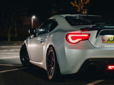 White sports car with red tail lights illuminated parked in car park at night with street light and UK license plate visible
