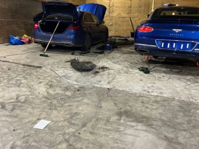 Blue Mercedes and blue Bentley luxury cars with open hood and tools inside industrial workshop with concrete floor and brick walls under industrial lighting
