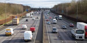 Multiple vehicles on multi-lane highway with yellow emergency response vehicle, white vans and trucks, cars in motion with bare trees and overcast sky