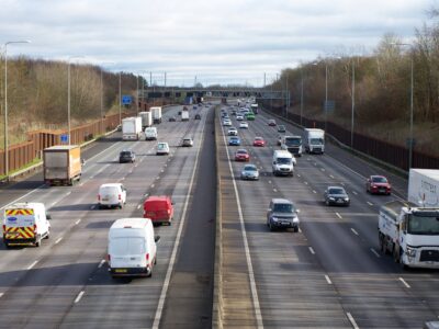 Multiple vehicles on multi-lane highway with yellow emergency response vehicle, white vans and trucks, cars in motion with bare trees and overcast sky