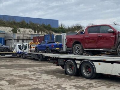Red Hilux recovery truck on flatbed with yellow police vehicle and industrial recovery yard with shipping containers and equipment visible