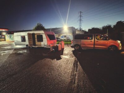 Orange stolen Toyota recovered vehicle parked at night in industrial recovery yard with caravans, facility lighting and power lines visible