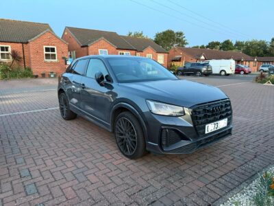 Dark grey Audi Q2 SUV recovered and parked on paved driveway in residential area with brick houses, trees and clear blue sky background