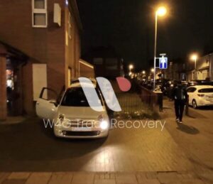 Recovered white car with red and white recovery stripe markings parked on nighttime urban street with street lights and brick buildings visible