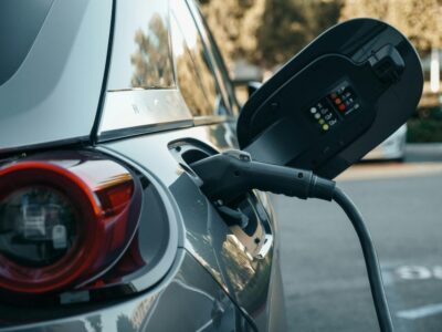 Close-up of black electric vehicle charging cable plugged into charging port with control unit buttons and vehicle red taillight visible