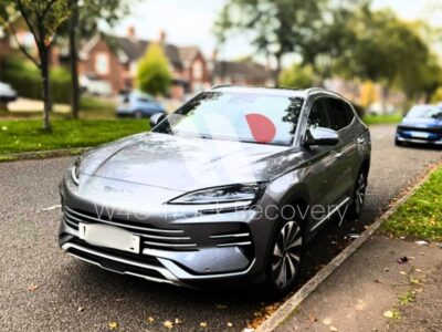 Silver electric vehicle with red recovery marking on windscreen parked on residential street with brick buildings and mature trees in background