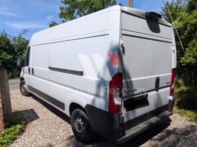 White and grey recovery work van with red tail lights parked in residential driveway with brick wall and green hedges under sunny daytime conditions