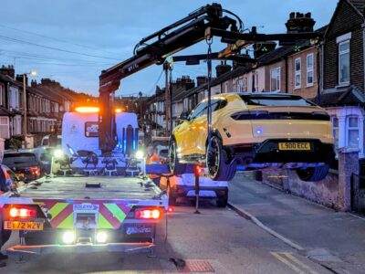 Yellow Lamborghini supercar being loaded onto recovery truck at night on busy urban street with emergency vehicles, street lights and brick buildings visible
