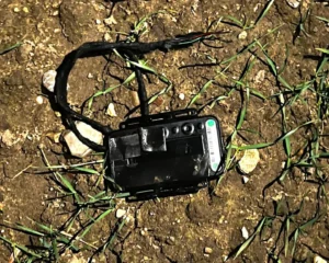 Black GPS car tracking device with white antenna and turquoise indicator light placed on brown soil ground with green grass sprouting around