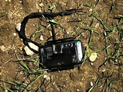 Black GPS car tracking device with white antenna and turquoise indicator light placed on brown soil ground with green grass sprouting around