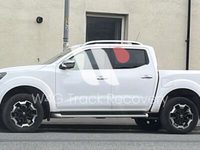 White Nissan Navara double-cab pickup truck parked on residential street in front of cream colored house with dark roof